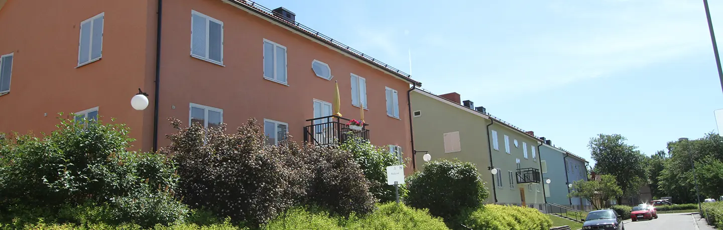 Tre tvåvåningshus med putsade fasader i orange, grön och blå. Grönska och villagata i förgrund.