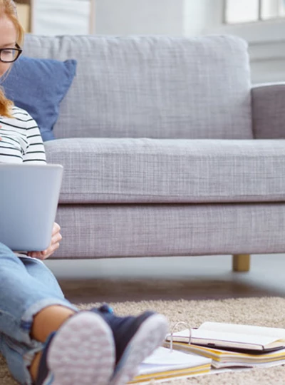 Tjej som sitter på golvet, lutad mot en soffa och studerar med en dator framför sig