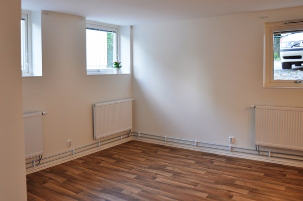 Ett rum med flera fönster, vita väggar och träfärgat golv.